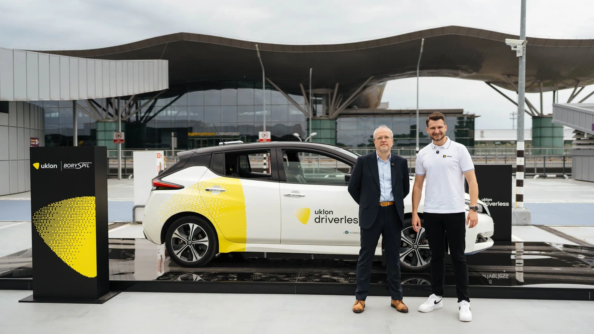 Презентація роботаксі Uklon driverless: двоє чоловіків біля білого автомобіля з жовтим брендуванням на виставковому майданчику.